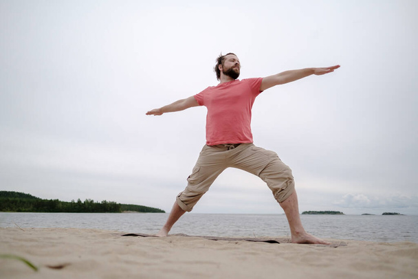Běloch cvičí jógu, cvičí virabhadrasanu, stojí na koberci, na písečné pláži u jezera. - Fotografie, Obrázek