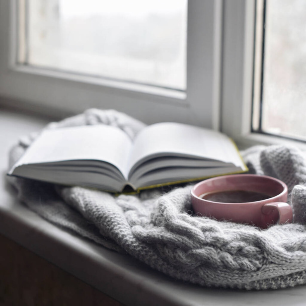 Cozy home still life: cup of hot coffee and opened book with warm plaid on windowsill against snow landscape outside. Winter holidays and Christmas time concept, free copy space. - Photo, Image