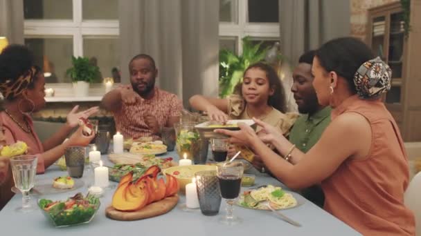 Mittlere PAN-Aufnahme einer großen, fröhlichen afro-amerikanischen Familie, die zu Hause gemeinsam zu Abend isst, plaudert und Teller austauscht, um besondere Anlässe zu feiern - Filmmaterial, Video