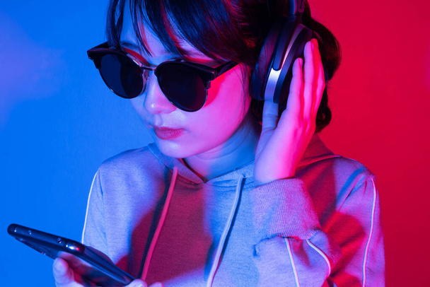 Nuori aasialainen nainen kuulokkeissa kuuntelee musiikkia tai pelaa peliä älypuhelimella neonvalolla taustalla. - Valokuva, kuva