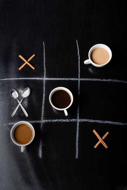 vista superior del juego tic tac toe con cucharas, palitos de canela y tazas de café en negro - Foto, Imagen
