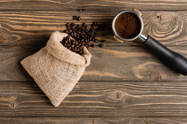 vrchní pohled na pytel s fazolemi v blízkosti kovového portafiltru s čerstvou mletou kávou na dřevěném povrchu - Fotografie, Obrázek