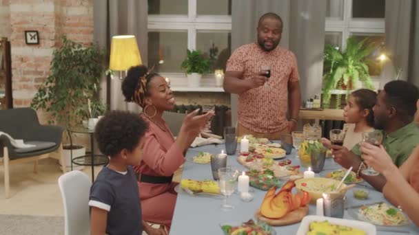 Mediana toma de hombre afro-americano feliz de pie frente a la familia en la gran mesa de la cena haciendo tostadas y luego tintineo vasos juntos - Metraje, vídeo