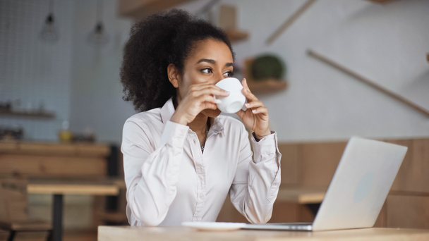 mujer afroamericana bebiendo café cerca del ordenador portátil en la cafetería - Foto, imagen
