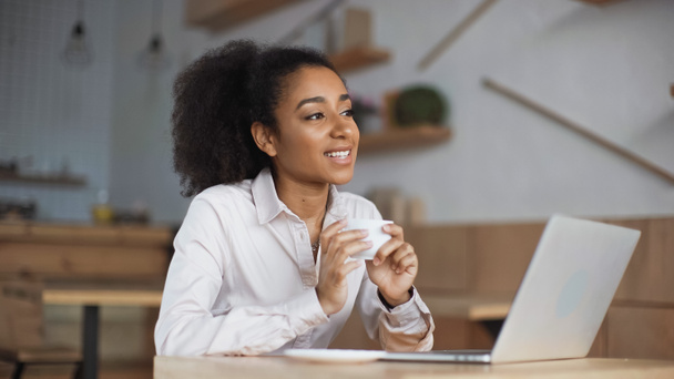 happy african american woman holding cup of coffee near laptop in cafe - Photo, Image