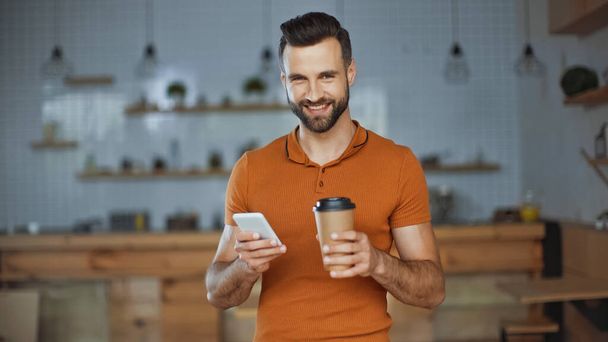 hombre sonriente usando teléfono inteligente y sosteniendo café para ir en la cafetería - Foto, Imagen