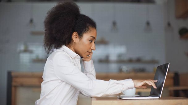 vista laterale della donna afro-americana che parla su smartphone e indica il computer portatile nel caffè  - Foto, immagini