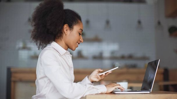boční pohled na usmívající se africké americké ženy držící smartphone a pomocí notebooku v kavárně  - Fotografie, Obrázek