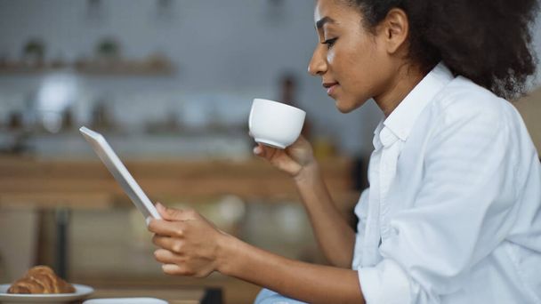 side view of african american woman using digital tablet while holding cup  - Photo, Image