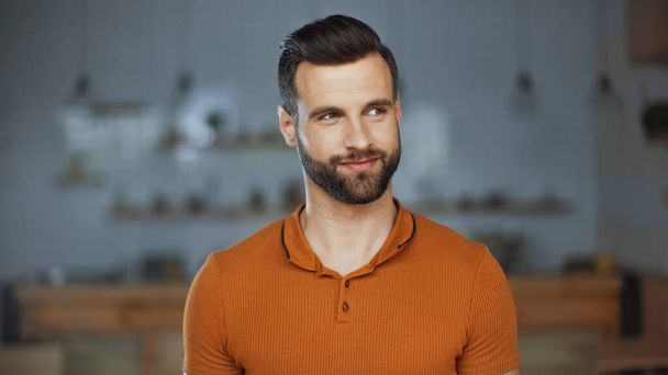 hombre barbudo en camiseta naranja mirando hacia otro lado y sonriendo en la cafetería - Foto, imagen
