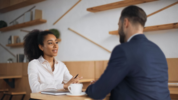 Африканская американская бизнесвумен разговаривает с размытым бизнесменом в костюме во время встречи в кафе  - Фото, изображение