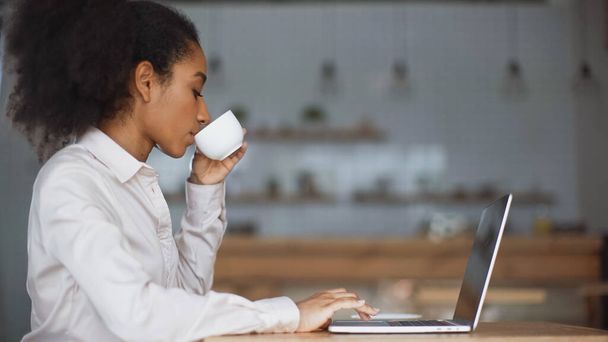 vista lateral de la mujer afroamericana bebiendo café y utilizando el ordenador portátil  - Foto, Imagen