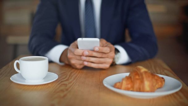 oříznutý pohled na podnikatele pomocí smartphonu v blízkosti croissantu a kávy v šálku na rozmazaném popředí  - Fotografie, Obrázek
