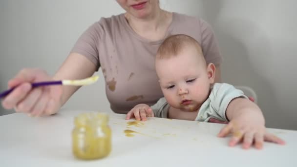 Malý novorozenec vtipný chlapeček se učí jíst zeleninové nebo ovocné pyré ze skleněné nádoby s lžičkou. Mladá matka pomáhá malému synovi jíst první jídlo. - Záběry, video