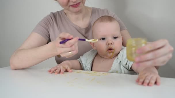 小さな新生児の面白い赤ちゃんの男の子はスプーンでガラス瓶から野菜や果物のピューレを食べることを学ぶ。幼い息子を助ける若い母親は最初の食べ物を食べる. - 映像、動画