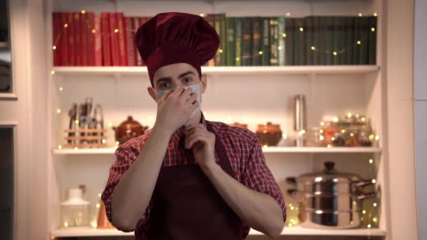 Cozinheiro sorridente no toque vermelho e avental está usando máscara facial e cruzando as mãos de pé em uma cozinha - Filmagem, Vídeo