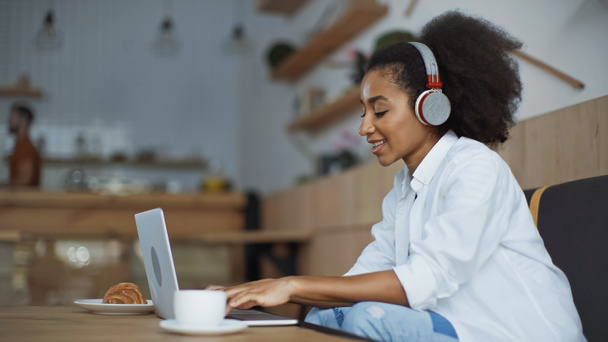 mujer de negocios afroamericana feliz en auriculares inalámbricos utilizando el ordenador portátil en la cafetería - Foto, imagen