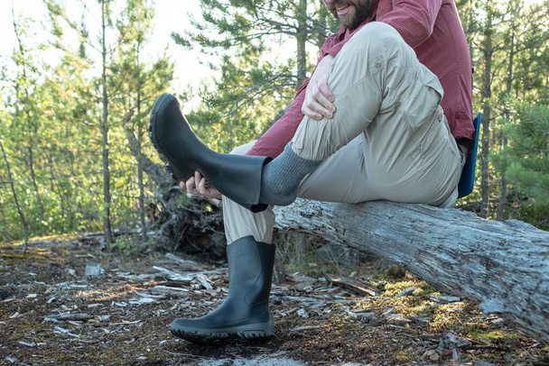 Человек в туристической одежде сидел на старом бревне в лесу, отказываясь от резинового ботинка, который стряхивает с него пыль. Концепция активного образа жизни. - Фото, изображение