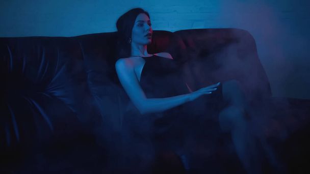 brunette woman in dress sitting on black couch on blue with smoke - 写真・画像