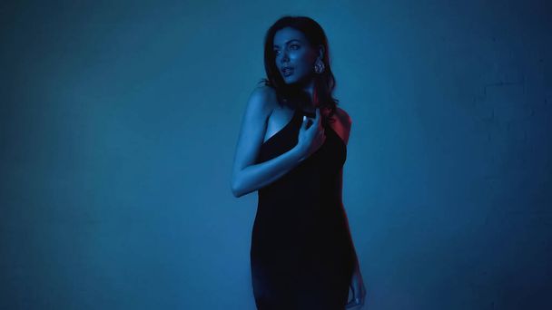 brunette woman in black slip dress looking away on blue background  - Фото, изображение