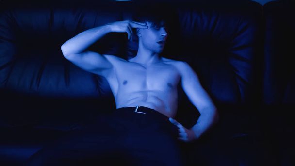 iluminación azul sobre el hombre sin camisa posando y haciendo gestos mientras descansa en un sofá negro  - Foto, imagen