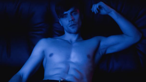 blue lighting on shirtless man in pants resting on black sofa  - 写真・画像