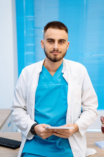 Serious young doctor in white scrubs poses in modern clinic office. Professional medic at working place. Closeup. - Photo, Image