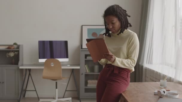 Afsluiting van jonge Afro-Amerikaanse vrouw met dreads in casual kleding in de woonkamer leunend op het bureaublad en werkend op digitale tablet - Video