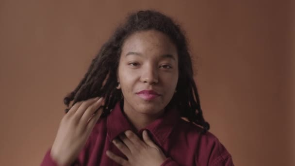 Foto mediana de una joven afroamericana con piel problemática y temores en la cabeza con camisa fijando su cuello mientras mira a la cámara y sonríe - Metraje, vídeo