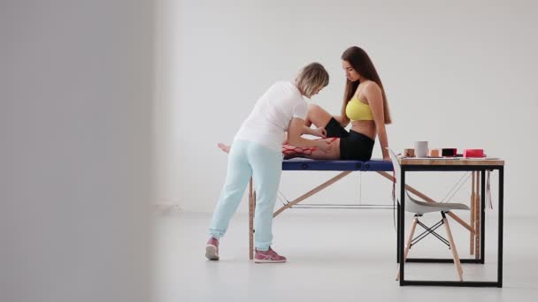 Женщина-физиотерапевт, накладывающая кинезио на ногу пациента в медицинском кабинете - Кадры, видео
