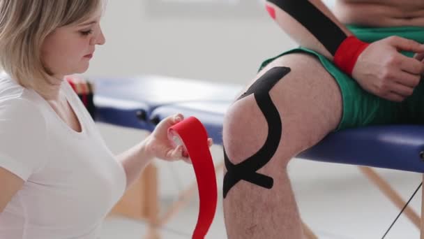 Fisioterapista che applica il nastro rosso di Kinesio ai pazienti gamba. Dottore tagliato pezzo di nastro adesivo dal rotolo nastro cinesiologia - Filmati, video