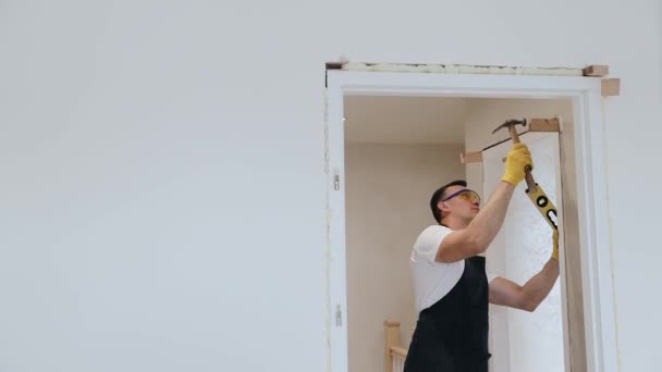 Αρσενικό ξυλουργός τεχνίτης σε εσωτερική ξύλινη πόρτα εγκατάσταση με σφυρί και το πνεύμα επίπεδο. Joiner συναρμολόγηση μια πόρτα ή jam μεταξύ δύο δωματίων σε νέο διαμέρισμα - Πλάνα, βίντεο