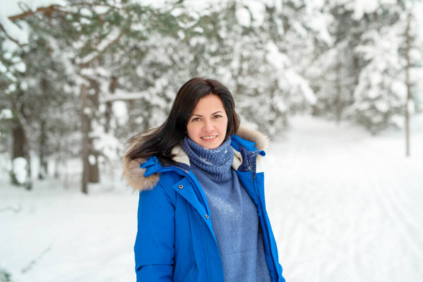 A young brunette woman in a blue jacket stands in the snow and watching into the camera in a winter forest. - Photo, Image