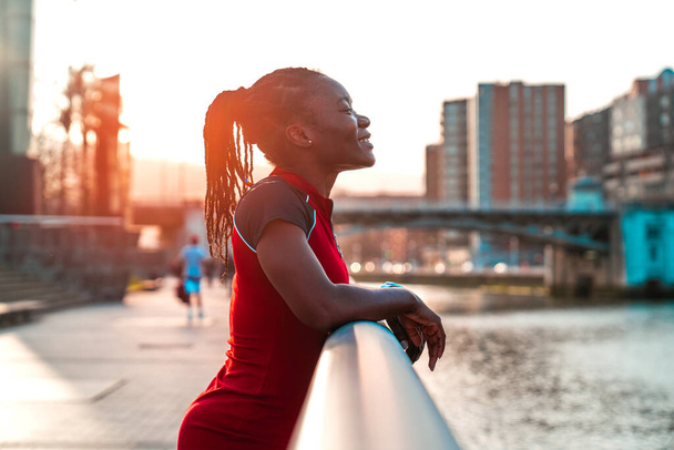 модель дівчина з чорною шкірою і волоссям в афро стилі позує посміхаючись тримаючись за перила мосту біля річки міста в красивому заході сонця з теплими кольорами в червоній сукні
 - Фото, зображення