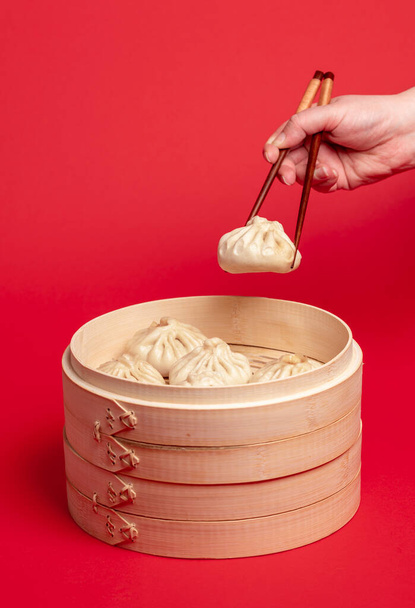 Woman taking a dumpling from the bamboo steamer with the chopsticks. Freshly steamed baozi dumplings in a wooden steamer isolated on a red colored background. - Photo, Image