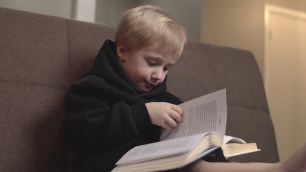 Aranyos kisfiú fekete kapucnis pulcsiban ül a kanapén és könyveket olvas. A kisfiú lapozgat a kanapén - Felvétel, videó