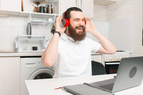 Чарівний молодий бородатий чоловік слухає музику на навушниках на кухні перед ноутбуком
. - Фото, зображення