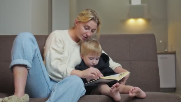 Cute little boy in black hoodie sits on couch with his mother and reads book - Footage, Video