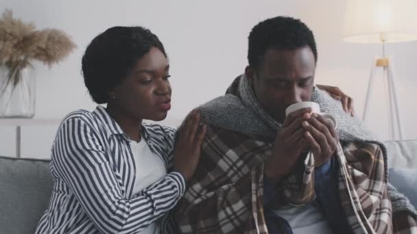 Opieka rodzinna. Kochająca afrykańska żona wspierająca swojego chorego męża, obejmująca go, mężczyzna pijący leczniczą herbatę - Materiał filmowy, wideo