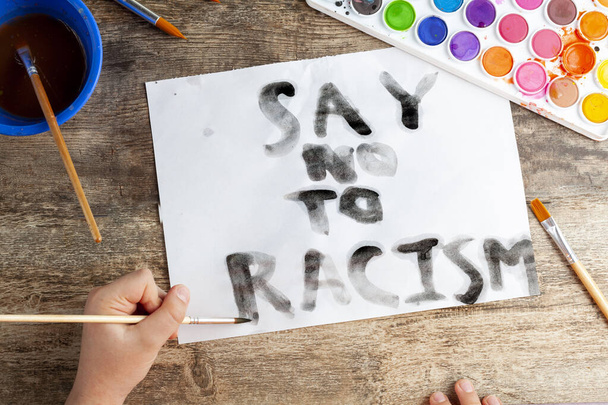 Enfant caucasien écrit "dire non au racisme" slogan en utilisant pinceau et aquarelle noire sur un livre blanc. Un enfant conscient des questions sociales telles que le racisme, l'inégalité et l'injustice concept - Photo, image