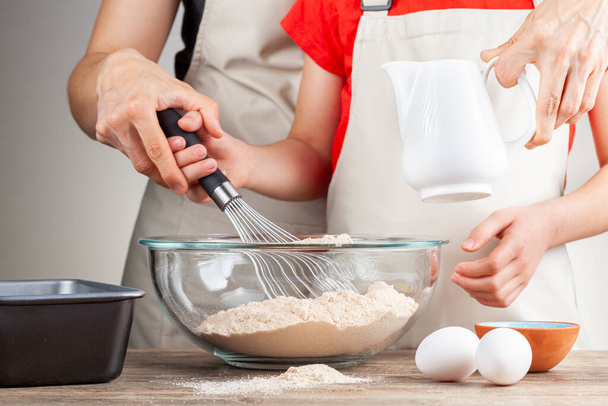 A mother is teaching her little daughter how to cook. They both wear aprons and together they mix a bowl of flour as part of a recipe. Mom and daughter cooking together concept. - Photo, Image