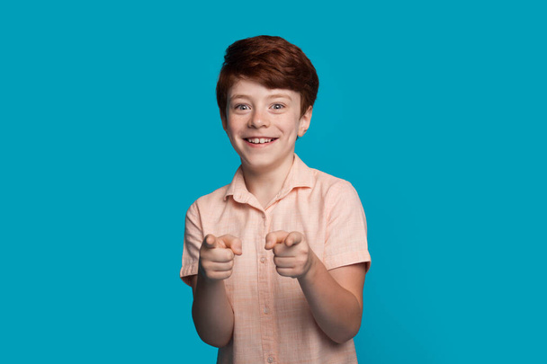 caucasien sourire garçon est geste à caméra avec doigt pointant sur un mur studio bleu avec espace libre - Photo, image