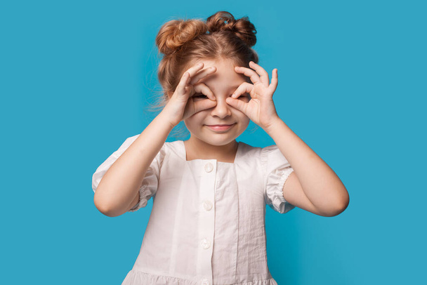 Caucasian girl is gesturing binoculars with fingers smiling at camera on a blue studio wall wearing a dress - Photo, Image