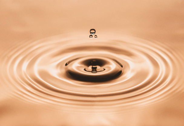 Waterdruppel druipt in een gouden poel en creëert rimpelingen en abstracte vorm van water. Drie druppels in camerafocus. Afbeelding bevat wat graan, geluid en bewegingsvervaging - Foto, afbeelding
