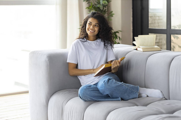 Ευτυχισμένη γυναίκα στο σπίτι κάθεται στον καναπέ κάθεται στον καναπέ διαβάζοντας βιβλίο. Όμορφη Αφροαμερικανή νεαρή γυναίκα χαλαρώνει διαβάζοντας το βιβλίο στο σπίτι στον καναπέ - Φωτογραφία, εικόνα
