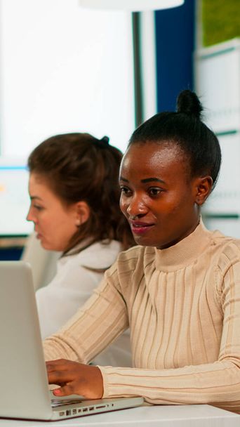 Iloinen afrikkalainen liiketoiminnan nainen kirjoittaa kannettavalla tietokoneella ja hymyilee istuu pöydällä - Valokuva, kuva