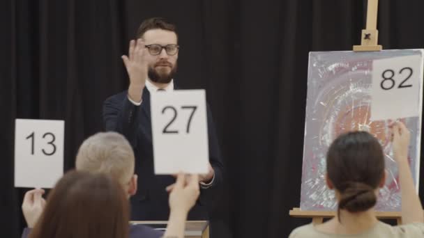 Мужчина-аукционист в очках и формальном костюме указывает на лопатки, поднятые участниками, и объявляет тендеры, продавая современную картину на аукционе искусств - Кадры, видео