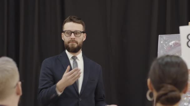 Professional bearded male auctioneer in eyeglasses and formal suit pointing at bidding paddles and calling out numbers while conducting art auction - Footage, Video
