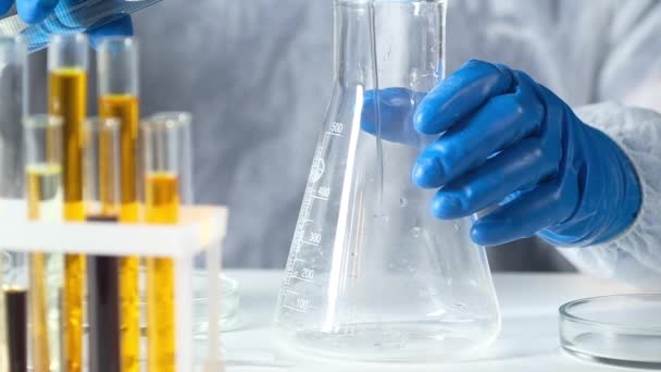 Chemicus in beschermend pak en handschoenen in modern lab giet heldere oplossing in grote glazen buis. Laboratoriumonderzoek en farmaceutische technologieën. Biologisch onderzoek in de farmacologische industrie - Video