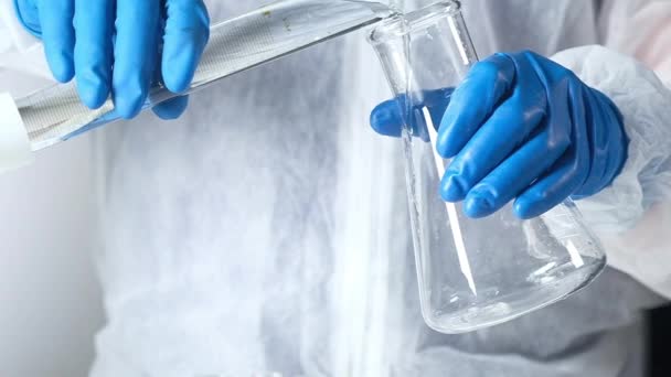 Il chimico in guanti protettivi il laboratorio moderno versa la soluzione limpida da becher in grande tubo di vetro. Tecnologie biochimiche e farmaceutiche di laboratorio. Ricerca biologica nell'industria farmacologica - Filmati, video